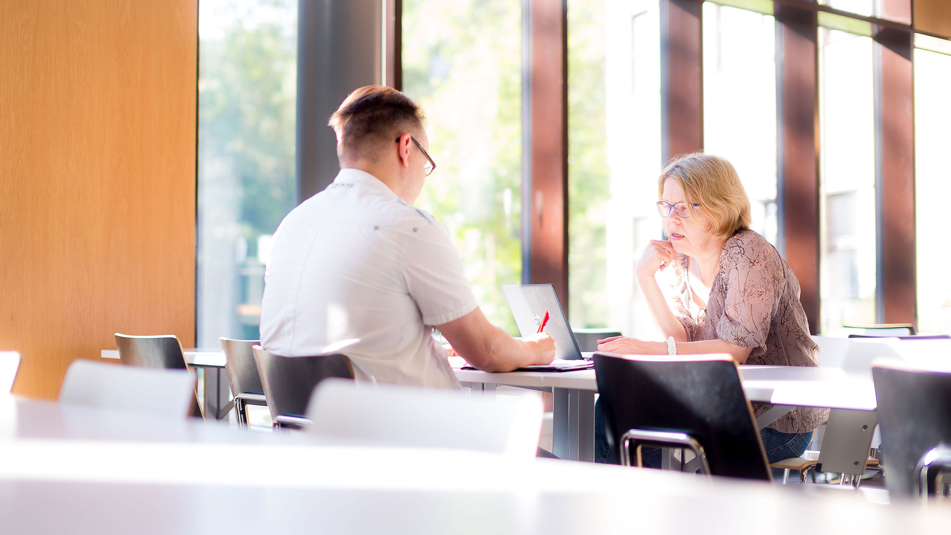 Two person sit in the opposite side of desk in a meeting. The other has a computer. From the big windows can be seen a park
