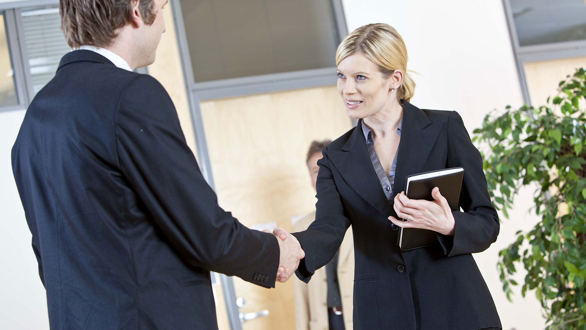 Blondhair woman with dark jacket handshakes dark-suited man