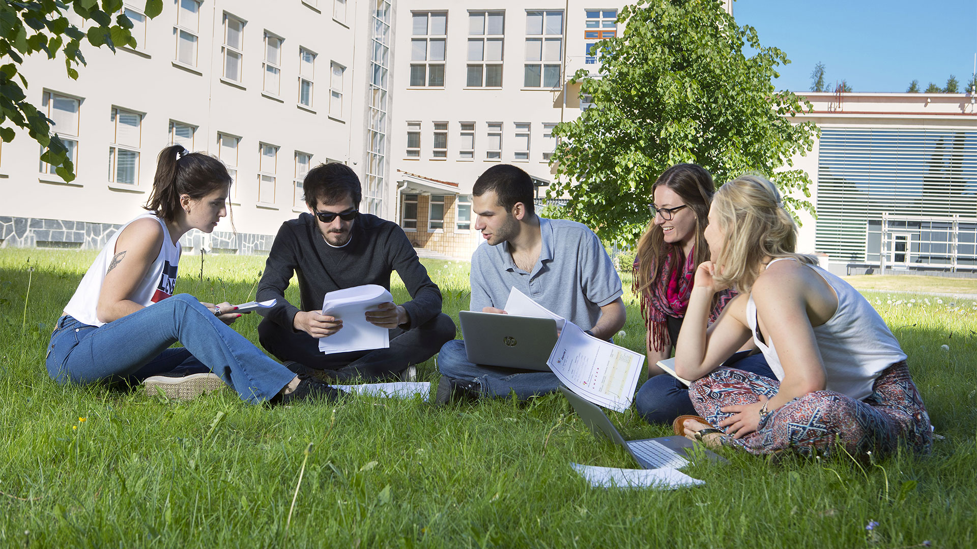 Students sitting on the grass with laptops. Sun is shining and in the back can be seen campus and trees