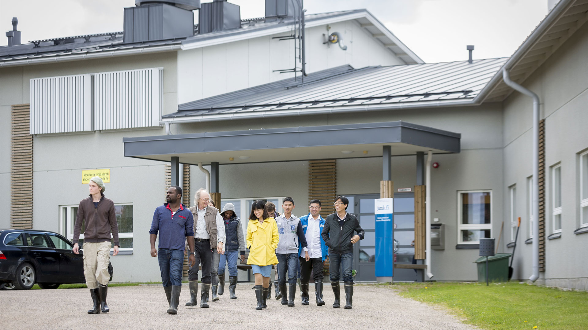 Group of students walks in rubber boots in front of grey buildings door towards the photographer