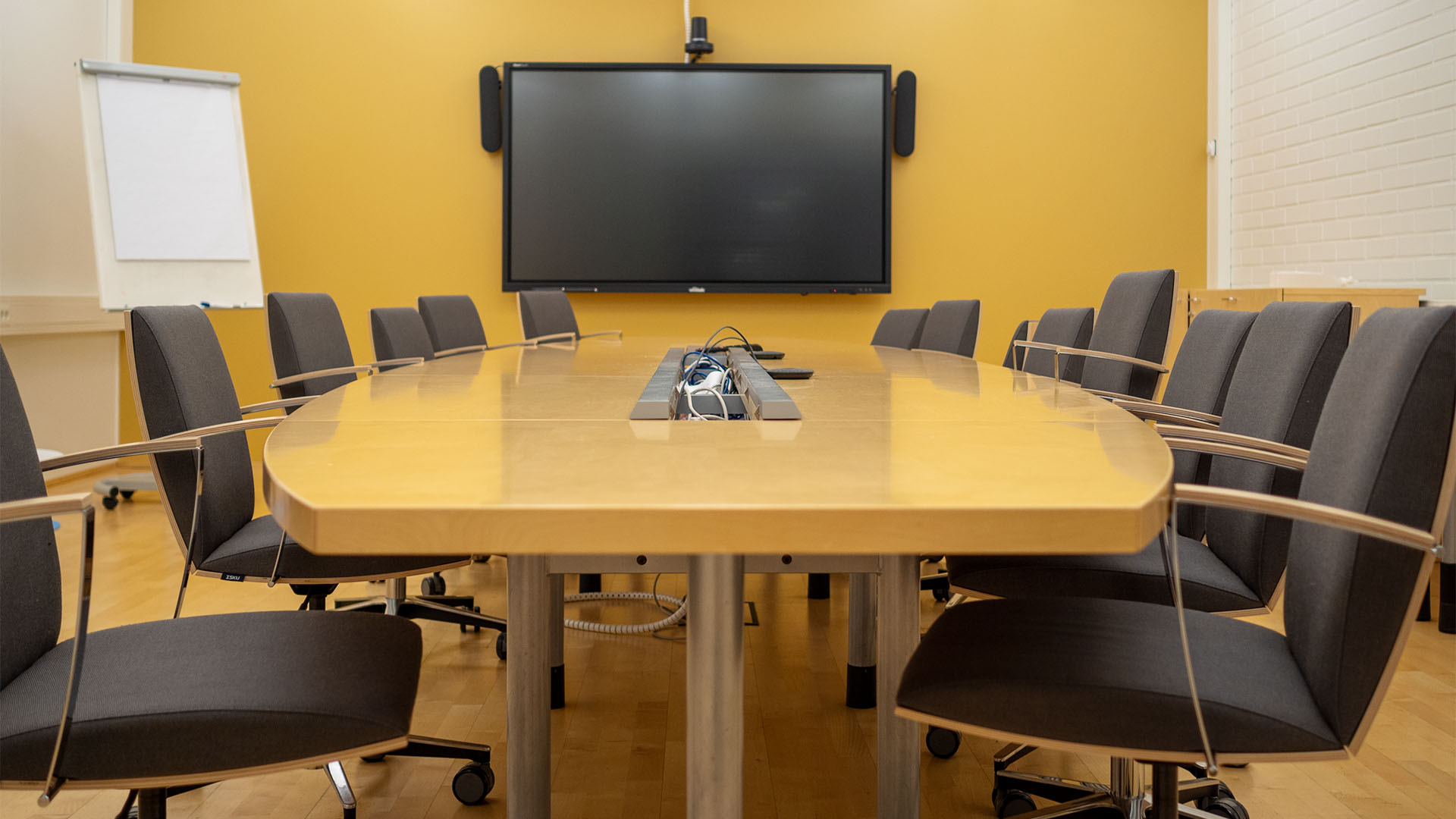 Neuvottelupöytä edestä päin. Takana seinällä näkyy näyttö, pyödän ympärillä tuoleja.