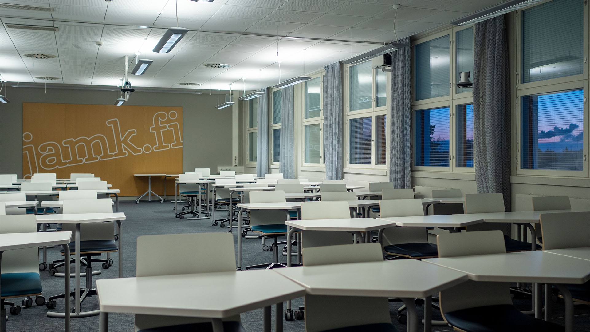 White desks and chairs in a rows. In the back there´s a wooden decorative wall. In the windows you can see sunrise.