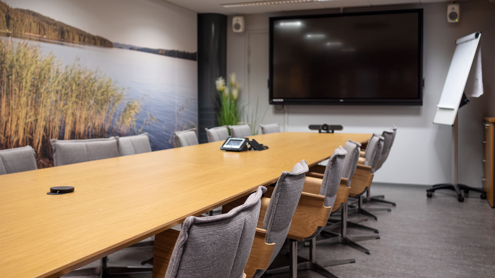 Neuvottelupöydän ympärillä tuoleja. Seinällä näkyyy maisematapetti järvikuvalla, toiselal seinällä iso näyttö.