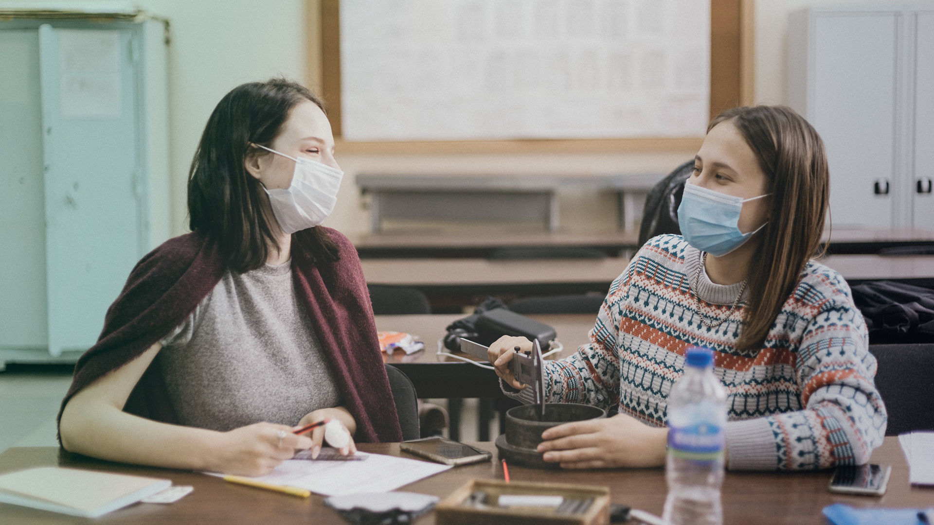 kaksi opiskelijaa katsoo toisiaan pöydän äärellä nenä-suu-suojaimet kasvoillaan