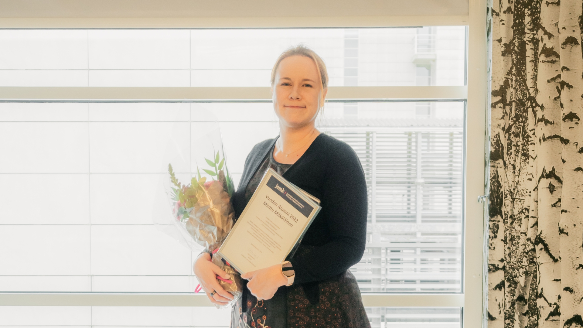 Nainen katsoo kameraan kunniakirjan ja kukkien kanssa