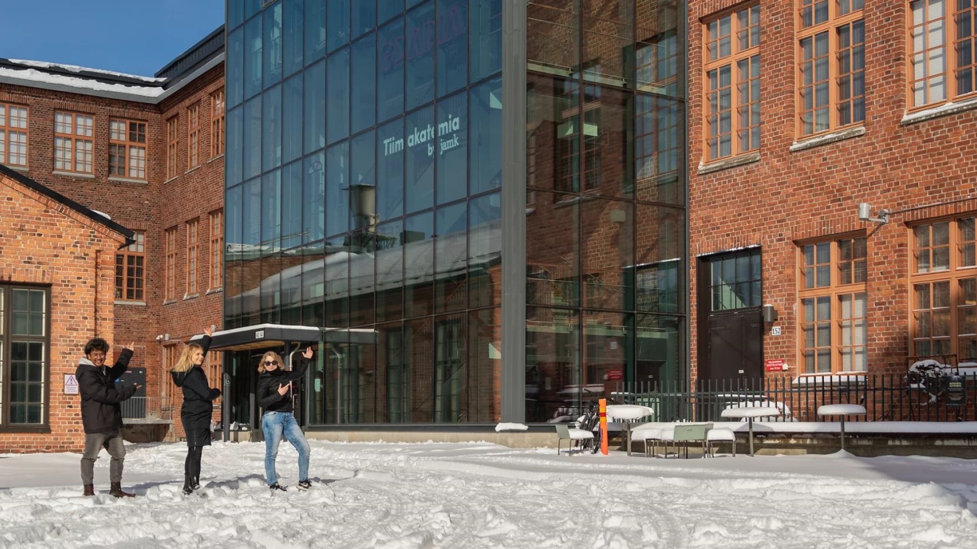 Tiimiakatemian Kankaan toimipisteen julkisivu ja Tiimiakatemian opiskelijoita talvisella pihalla osoittamassa Tiimiakatemian näyttävää valokylttiä rakennuksen lasiseinässä