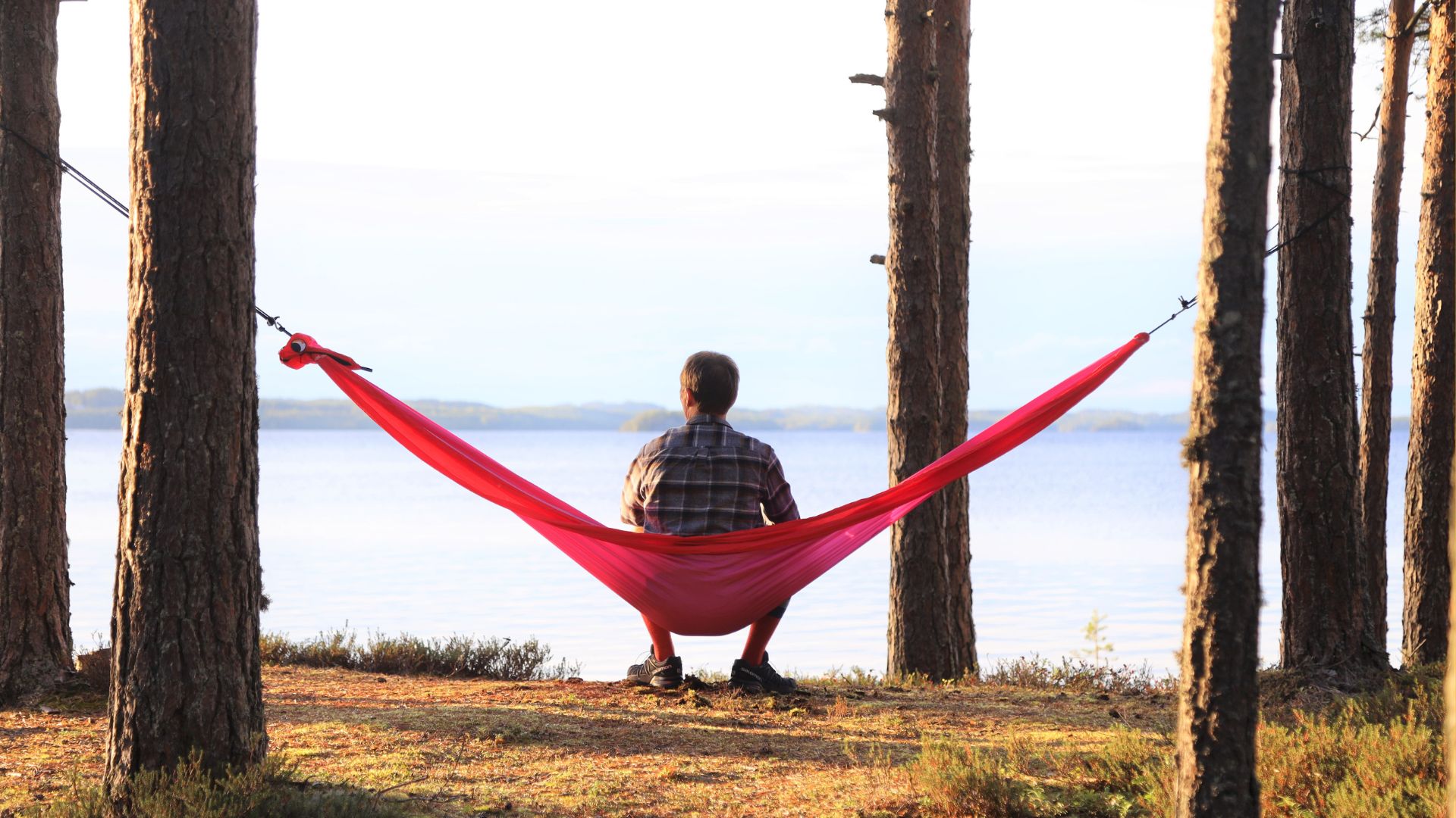 Henkilö istuu riippukeinussa katsoen järvimaisemaa selin kuvan katsojaan.