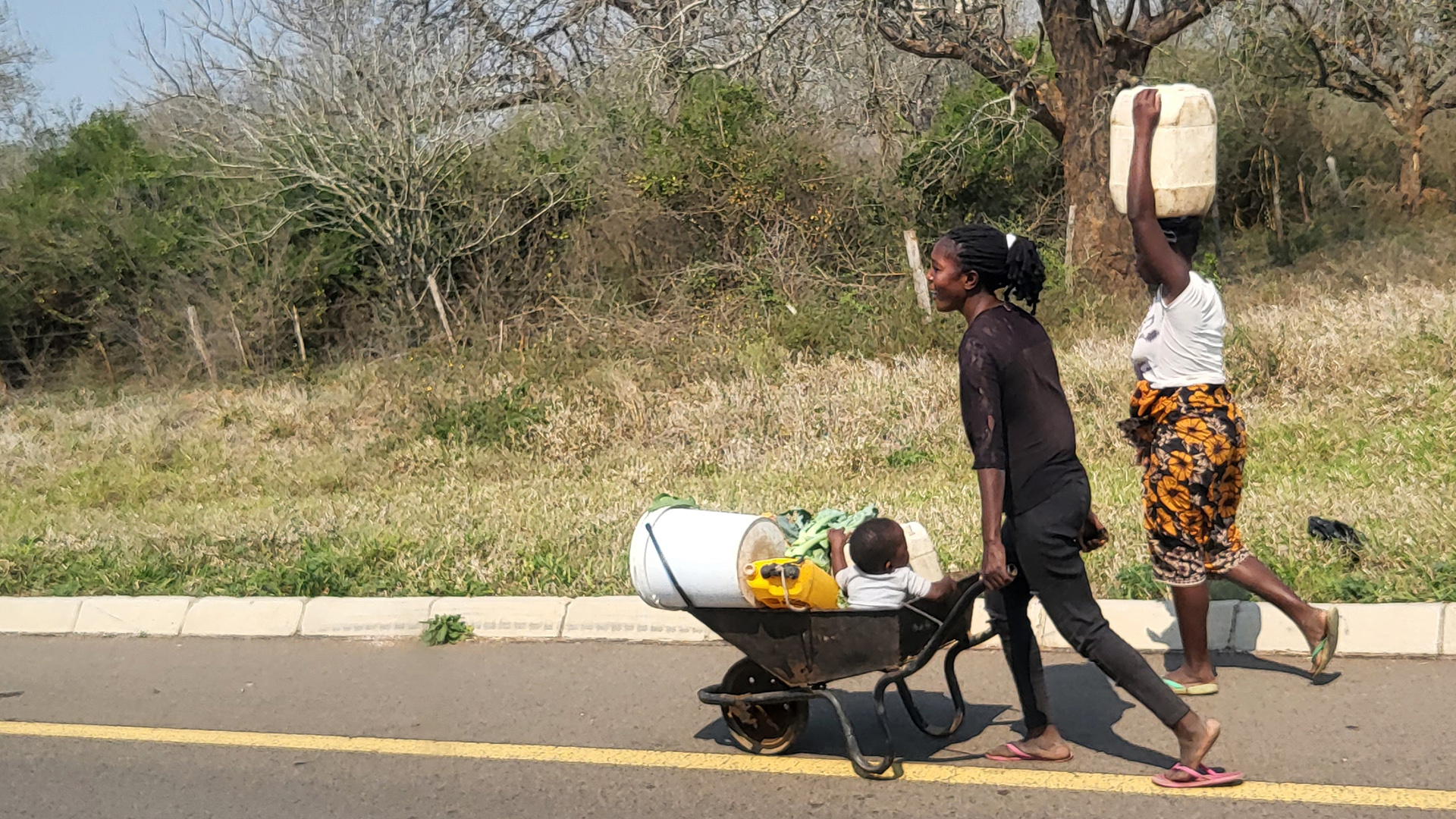 Women carrying water on their head and on a wheel barrow. A child sleeping on the wheel barrow.