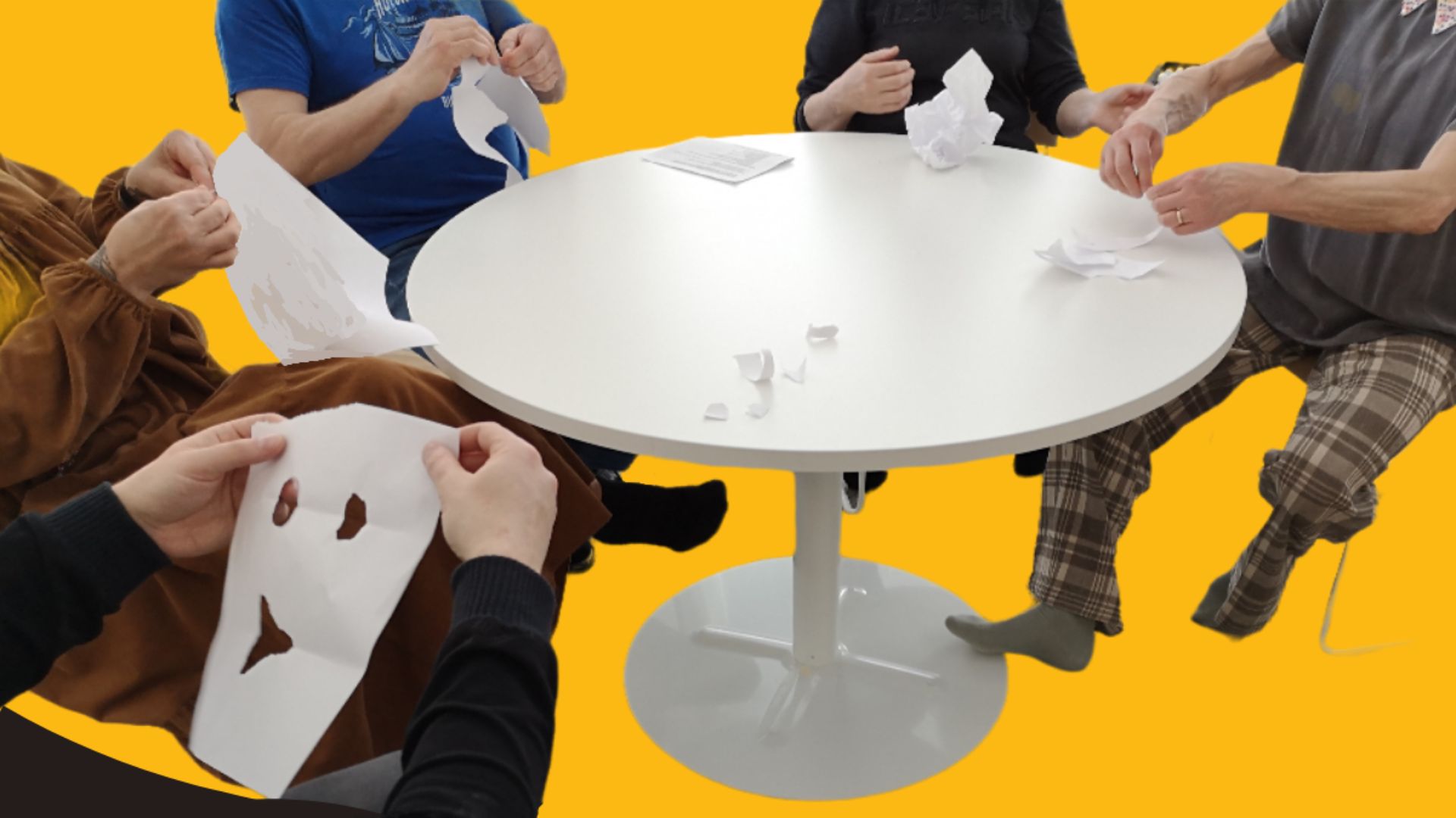 Ihmiset istuvat pyöreän pöydän ääressä ja muotoilevat paperiarkeista taidetta.