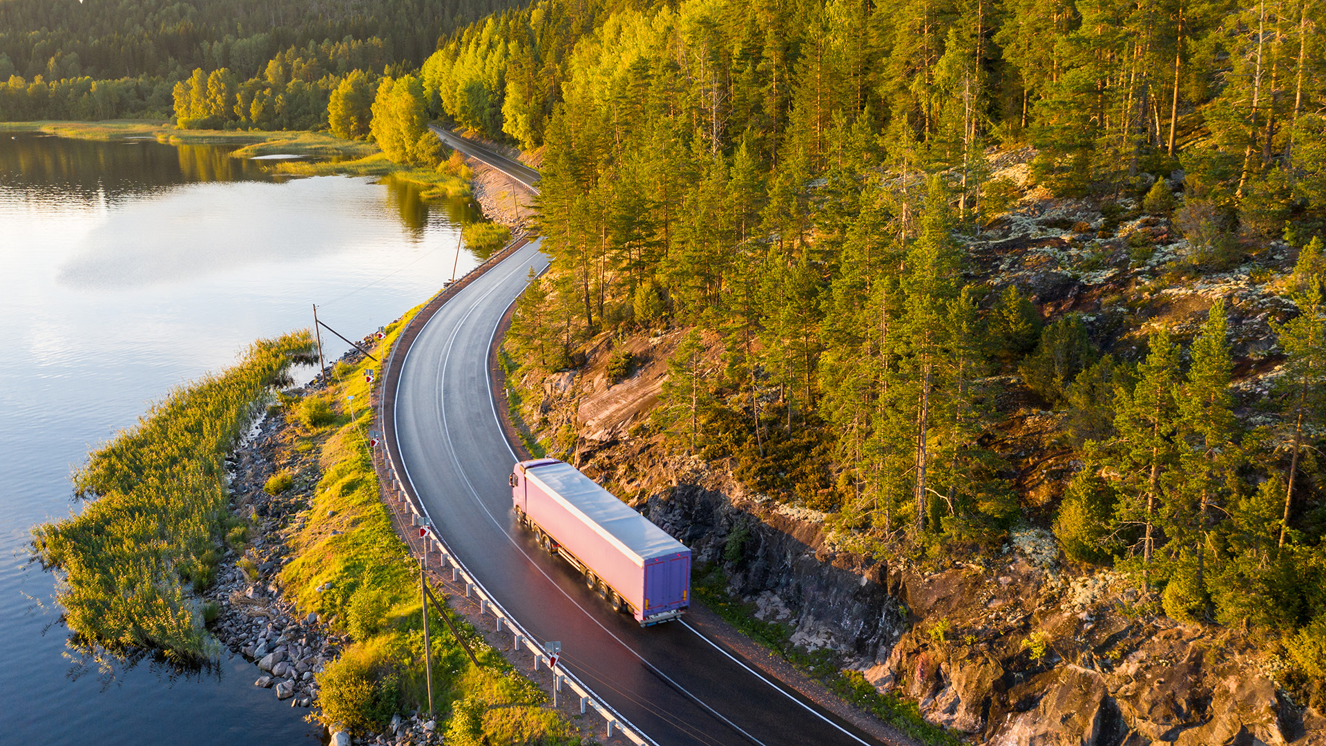 Rekka-auto ajaa tiellä järven rannassa, metsää.