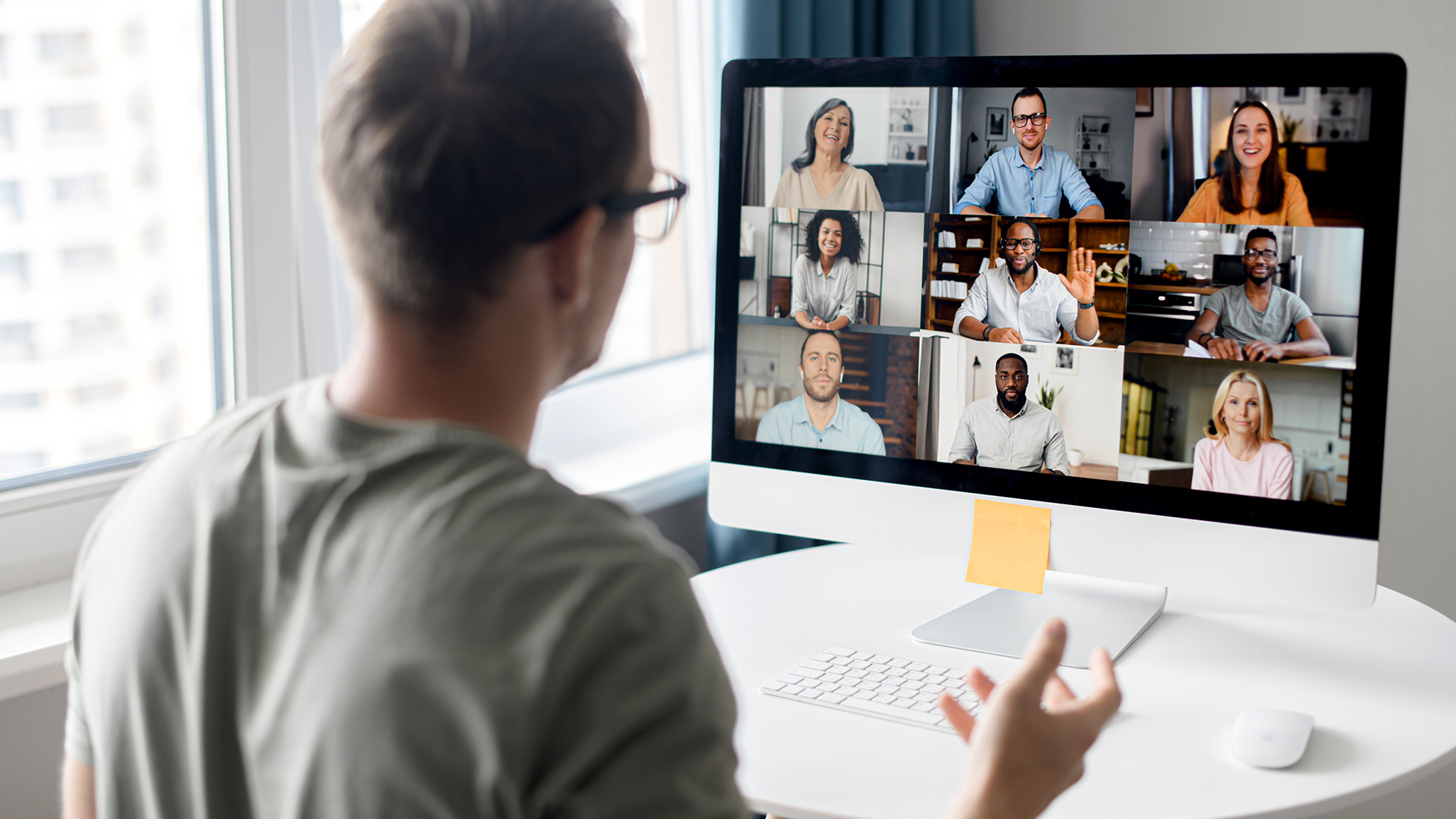 Takaa kuvattu henkilö osallistuu verkkokokoukseen, hänen toinen kätensä on kohotettu. Tietokoneen ruudulla näkyy 9 muun ihmisen kuvat.