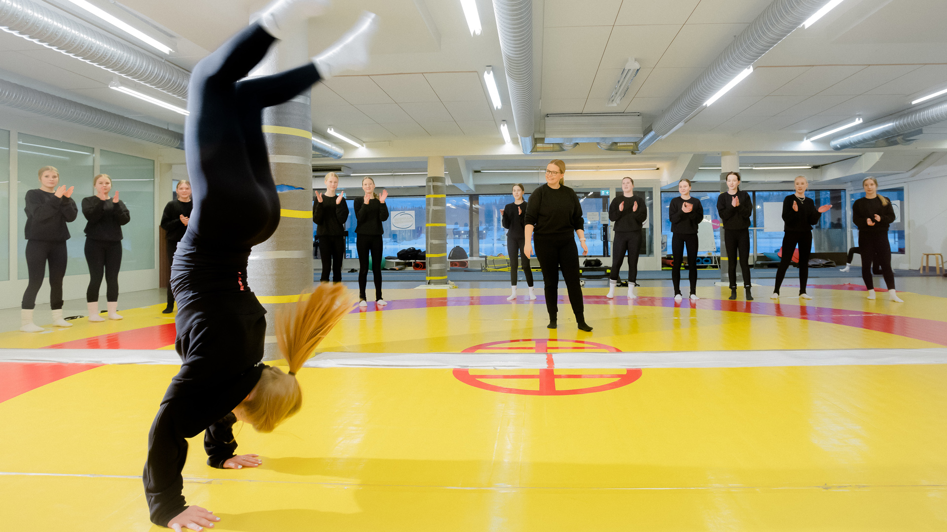 Tyttö seisoo käsillään Killerin treenitilassa muu joukkue ja valmentaja on taustalla.
