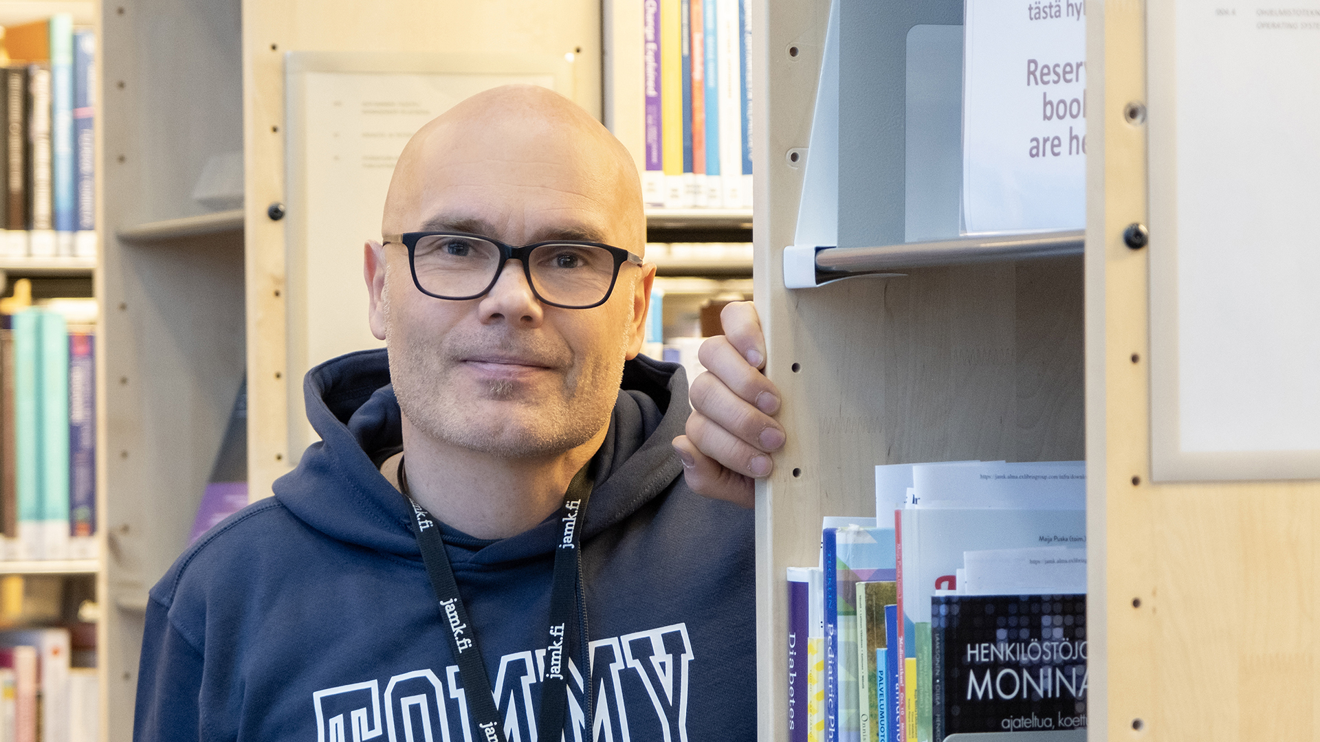 Mies seisoo kirjahyllyjen välissä ja katsoo hymyillen kameraan.