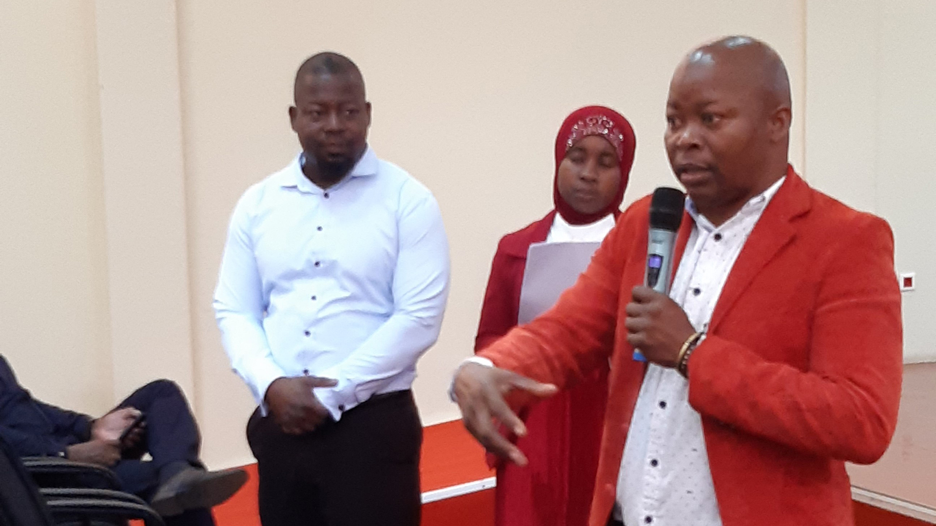 Projects key expert speaking in a microphone, two people standing next to him.