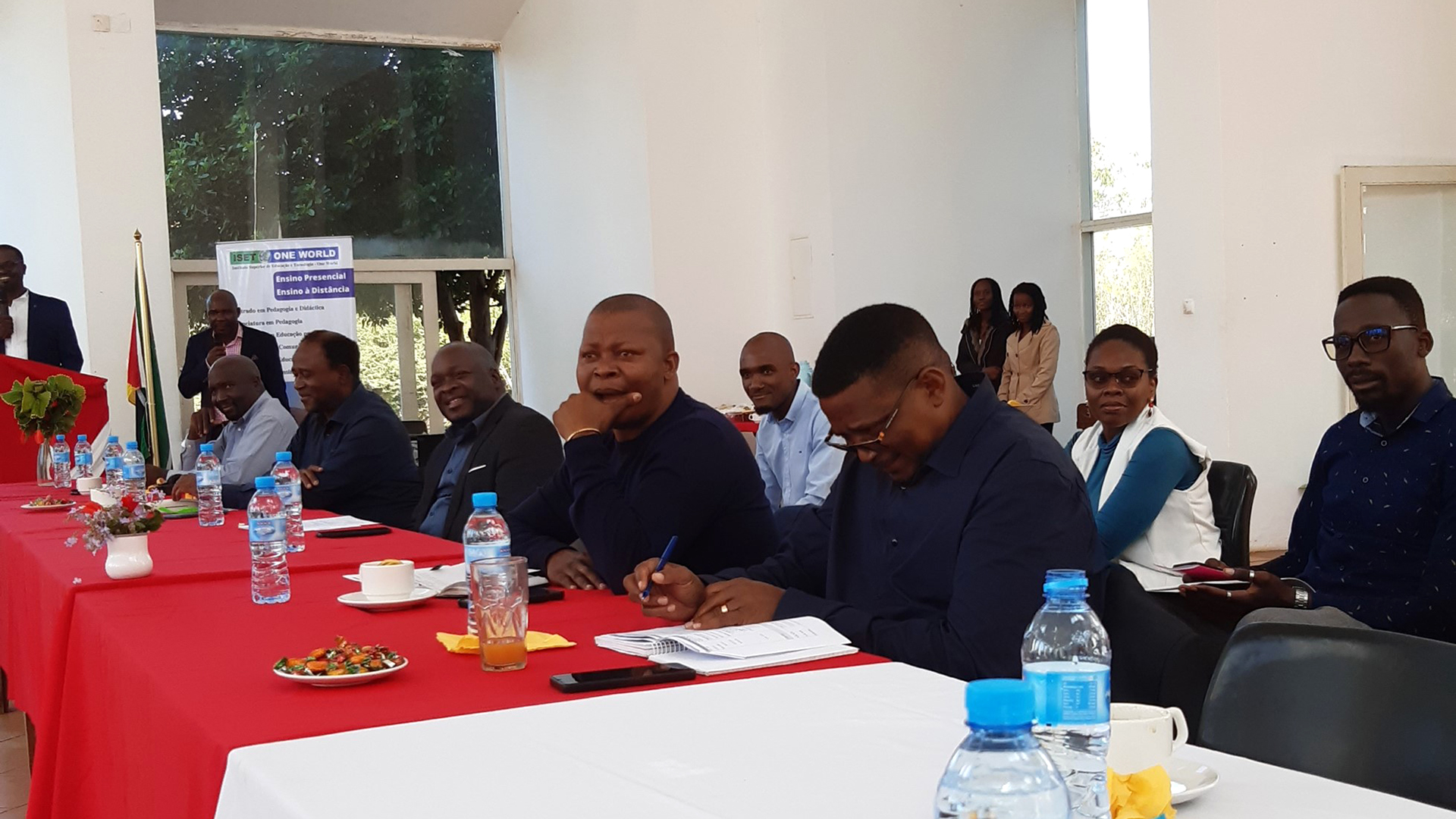 Eight people sitting by or behind a table which is covered by table clothes. Water bottles, notebooks, candies and flowers on the table.
