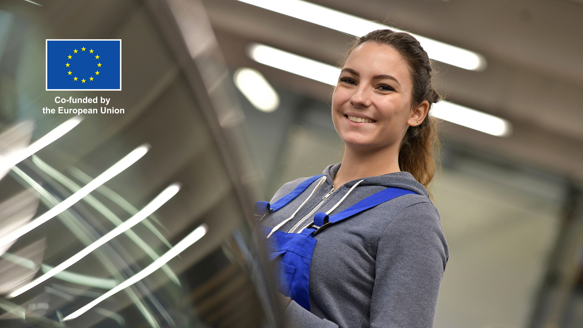 A trainee at car service, smiling next to a car wind shield. European union logo