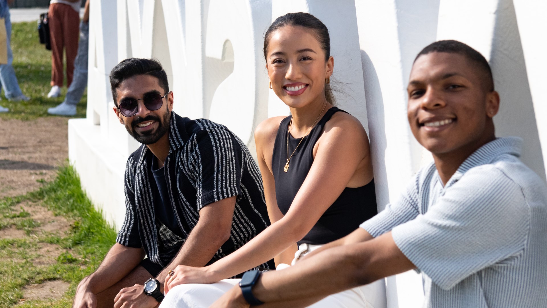 Three smiling students sitting outdoors in front of a public work of arts looking into the camera