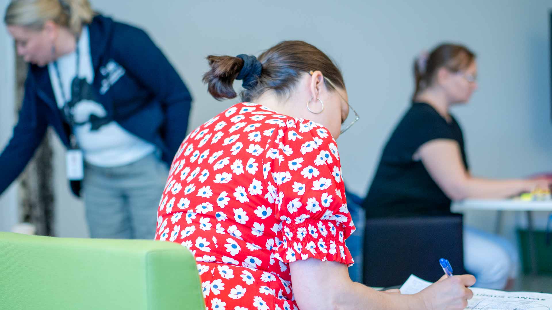 Punapaitainen nainen opiskelee pöydän ääressä, taustalla muita ihmisiä.