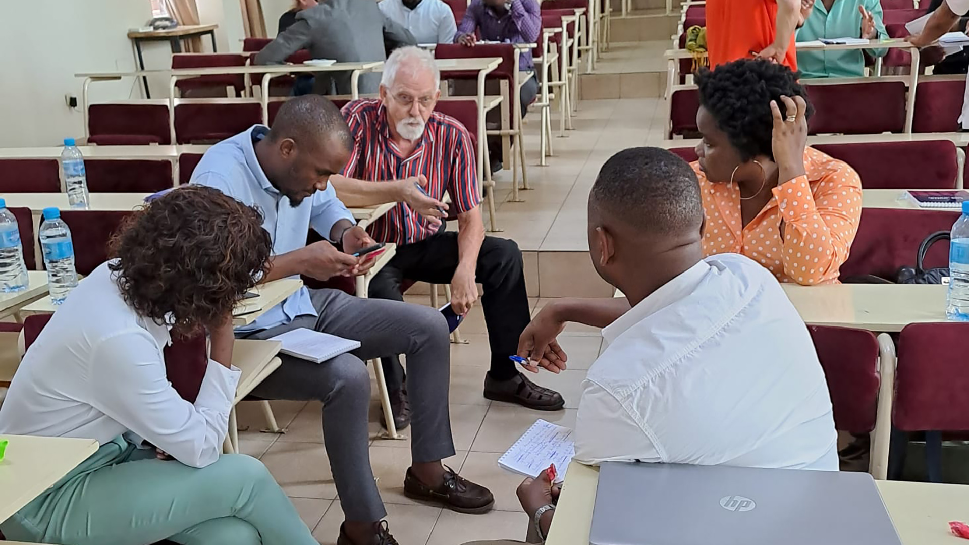 Five persons sitting in a group in between tables in class, and discussing.