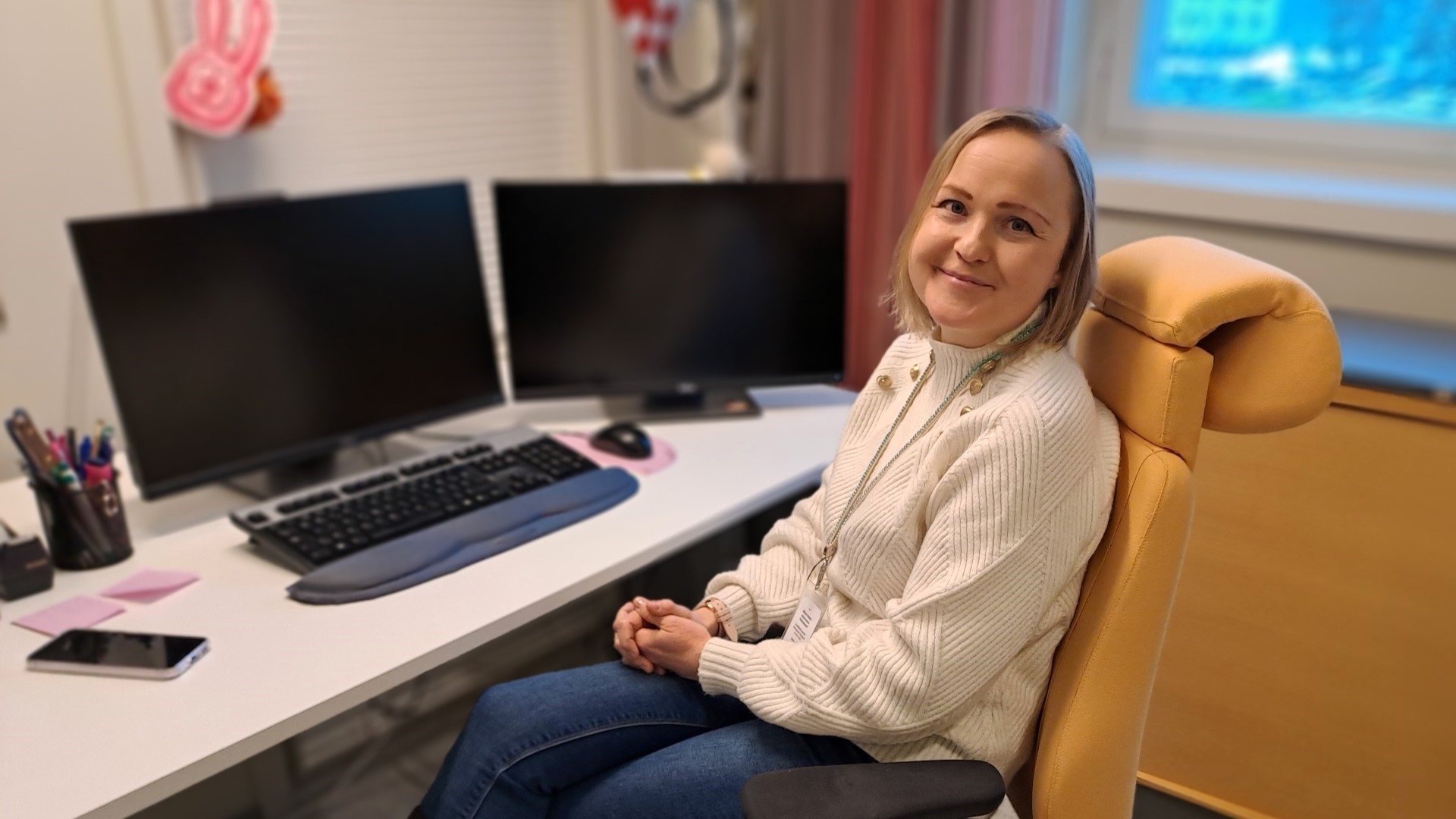 Soile Korhonen istuu työpöytänsä ääressä ja hymyilee suoraan kameraan. Pöydällä on tietokone, kaksi näyttöä ja ja puhelin.