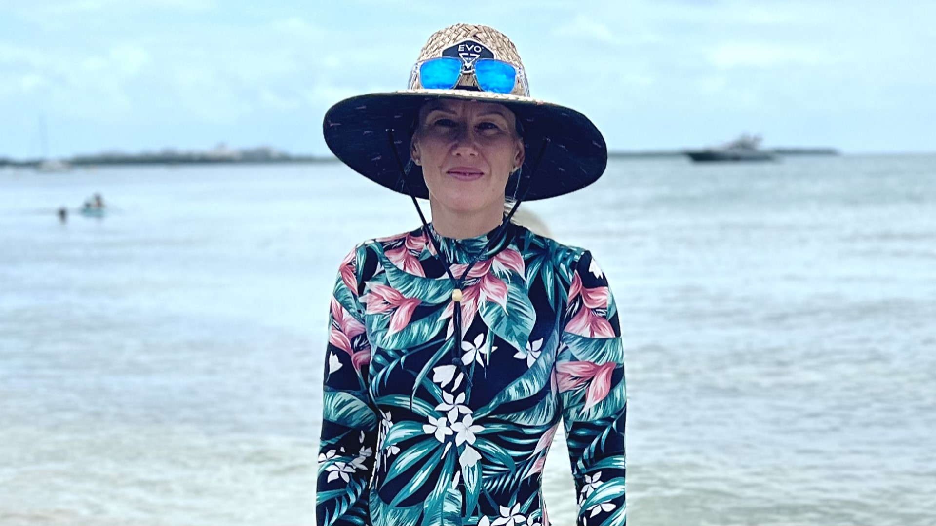 Nainen seisoo rannalla lierihattu päässään ja katsoo kameraan