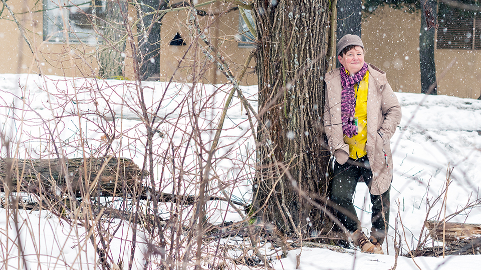 Nainen nojaa puuhun. Taustalla keltainen rakennus. Kuvan etualalla risukkoa.