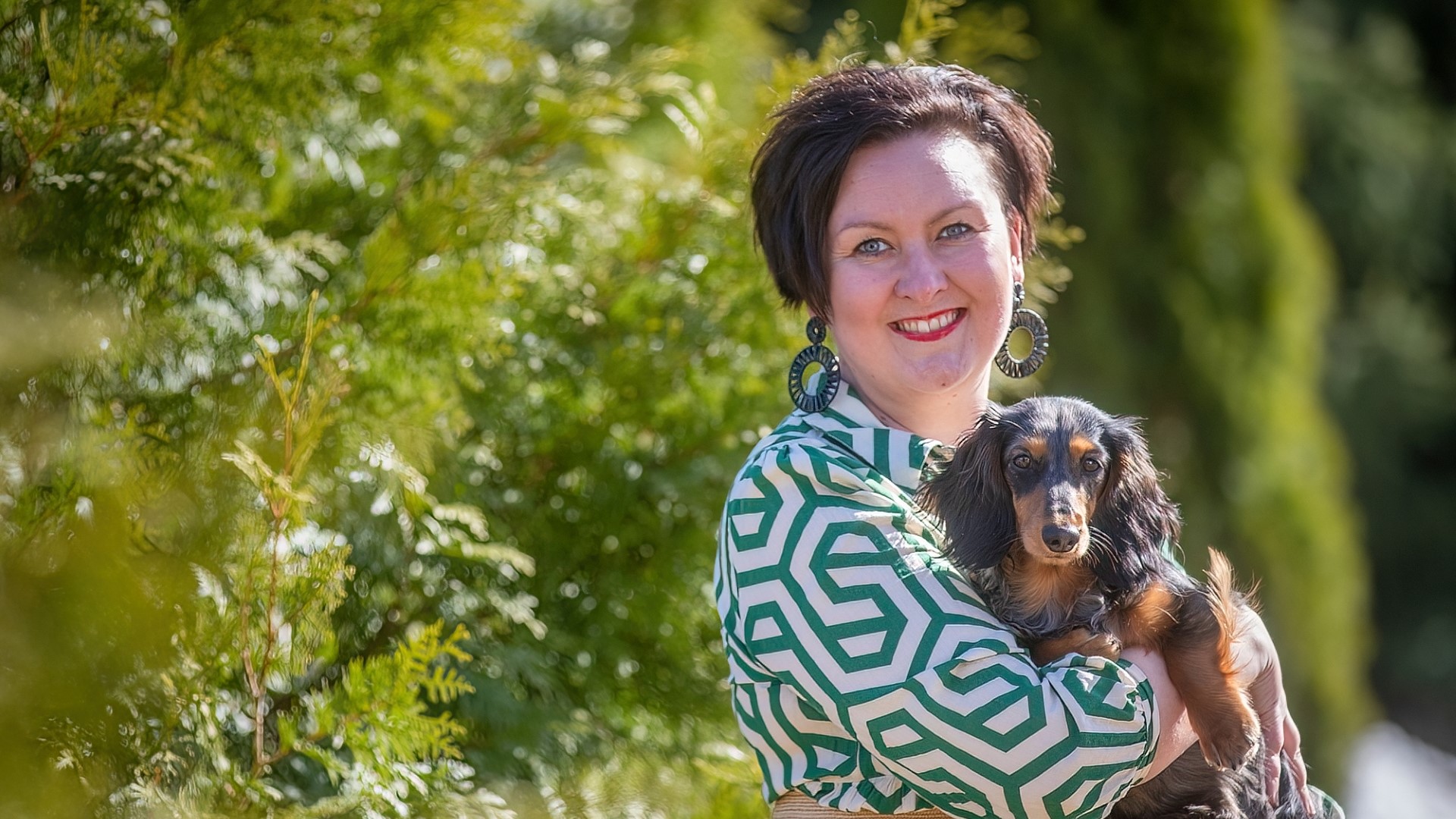 Laura Seppäsellä on mäykoira Lyyli sylissään. He molemmat katsovat kameraan. Aurinko paistaa.