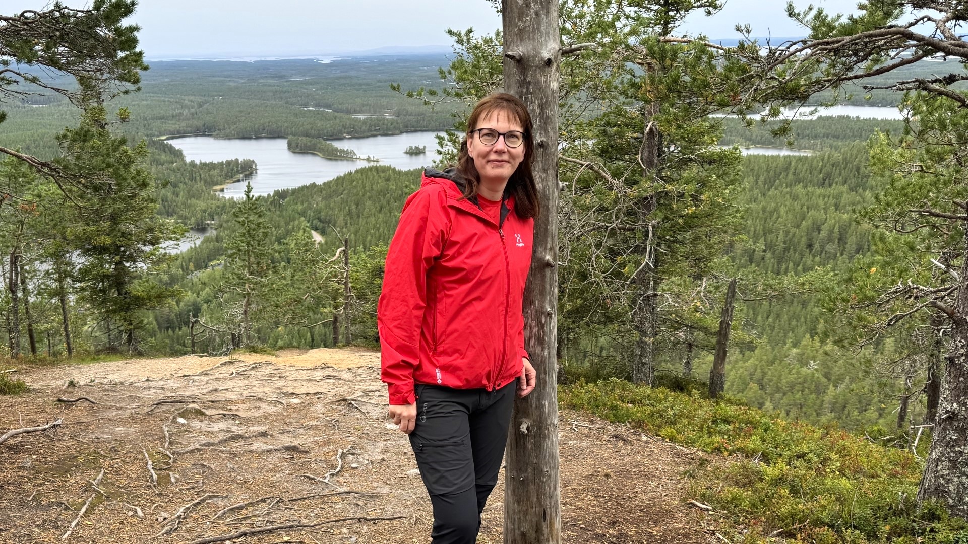 Sanna seisoo korkealla mäellä ja nojaa puuhun. Hän katsoo suoraan kameraan.