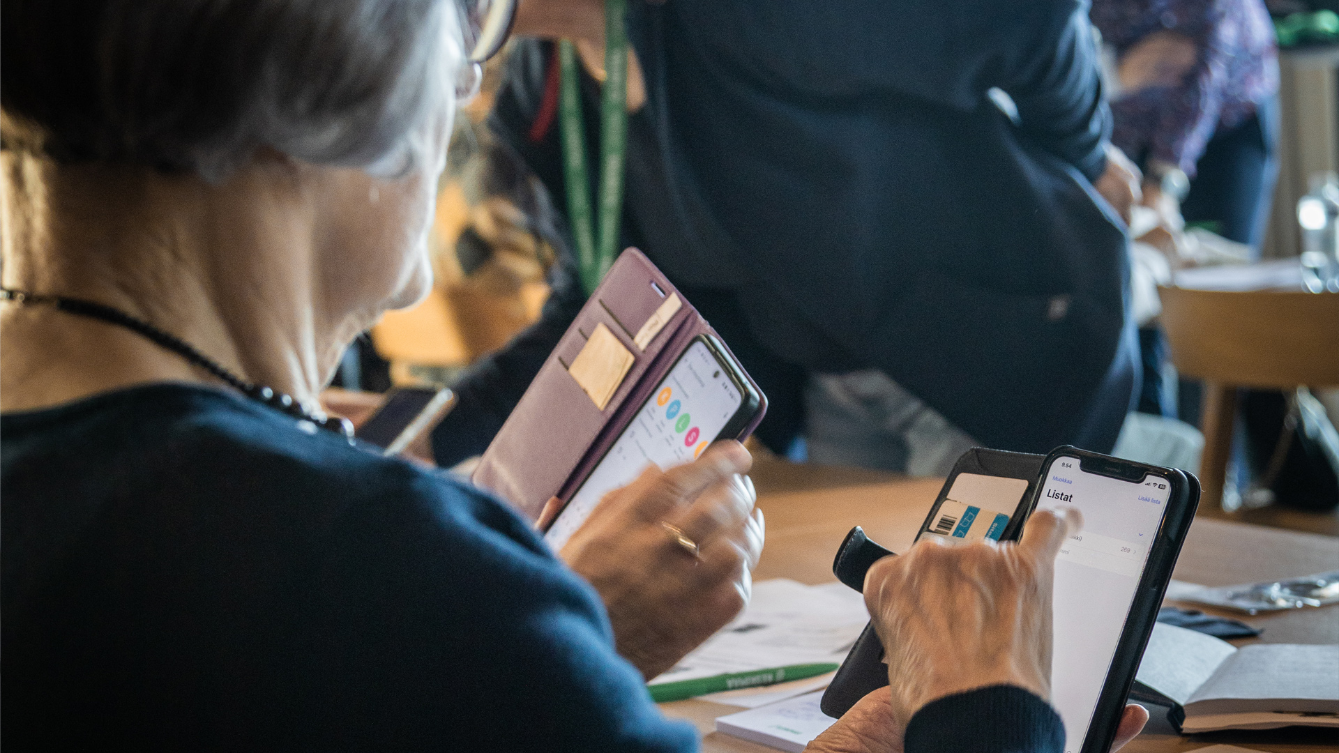 Kaksi ikäihmistä keskittyvät käyttämään älypuhelimia, taustalla kouluttajia..