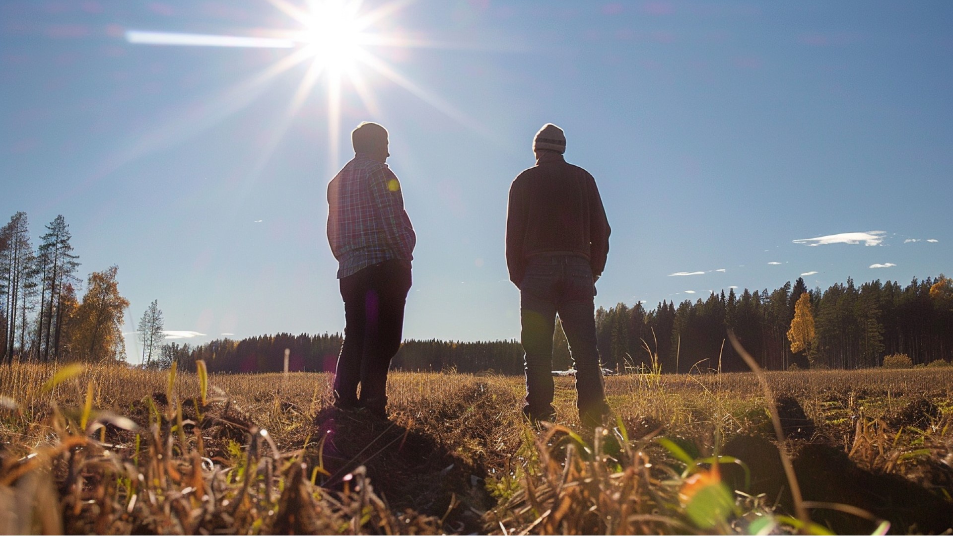 Kuvassa keski-ikäinen mies ja nuorempi pipopäinen mies katsovat vierekkäin seisten aukeaa peltomaisemaa auringon paistaessa.