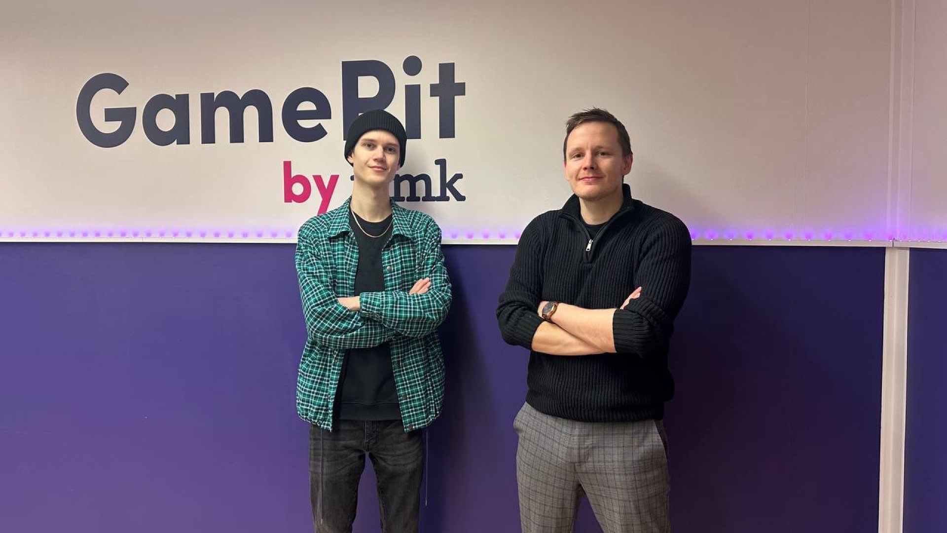Game Production Students Aaron Wirenius and Valtteri Yli-Olli standing in front of a GamePit by Jamk -logo, smiling, looking relaxed and confident