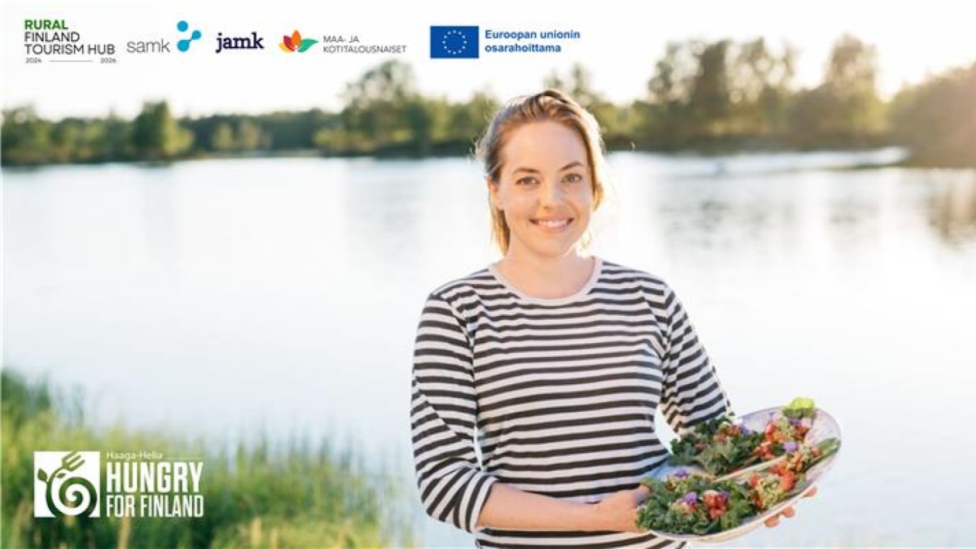 Iloinen henkilö matala kori käsissään suomalaisen järvimaiseman edustalla.