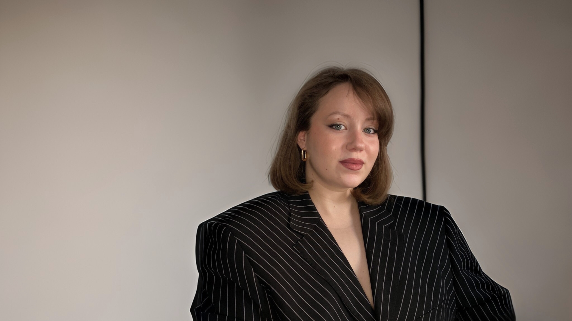 Tiia Manninen istuu harmaan seinän edessä ja katsoo suoraan kameraan.