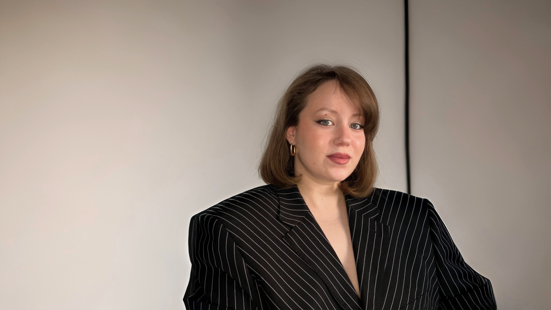 Tiia Manninen istuu harmaan seinän edessä ja katsoo suoraan kameraan.