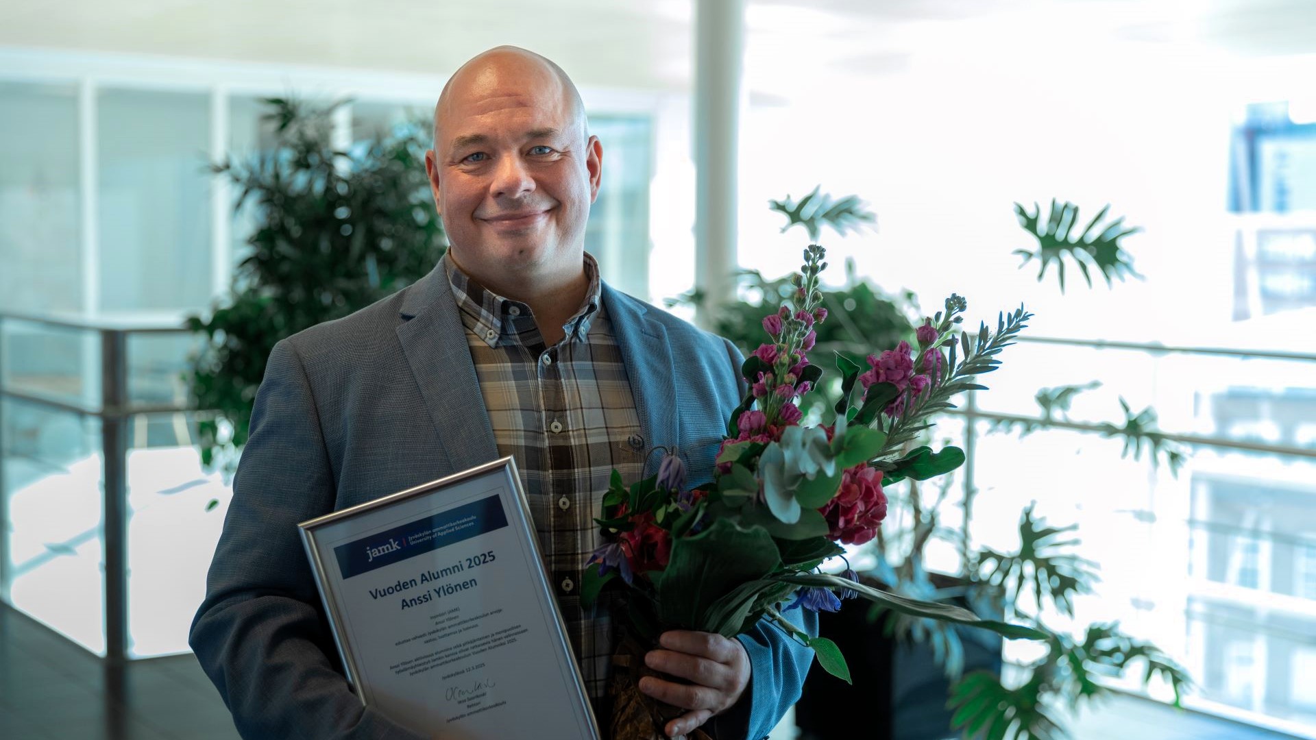 Anssi Ylönen katsoo hymyillen kohti kameraa. Hänellä on kädessään vuoden alumnin kunniakirja onnittelukukat.