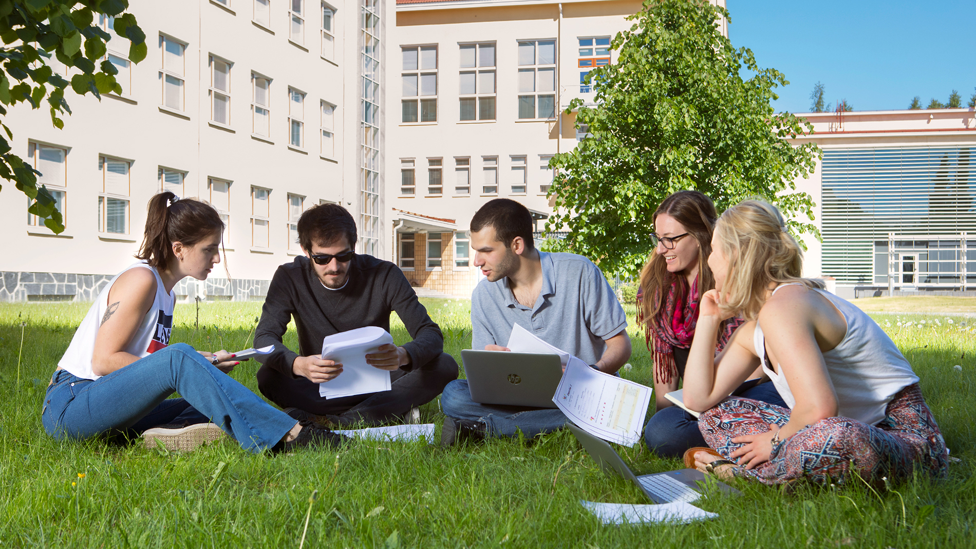Group of happy students sitting on the grass in front of the campus building.