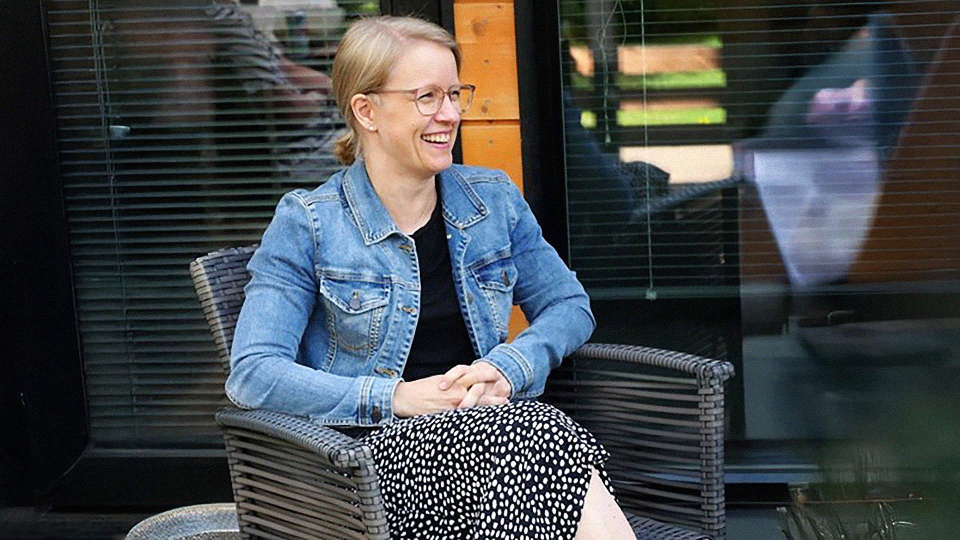 Hymyilevä henkilö farkkutakissa istuu puutarhatuolilla kädet sylissä.