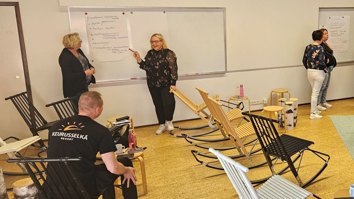 Kuvassa viisi henkilöä, joista yksi henkilö seuraa tapahtumia istuen. Neljä muuta henkilöä ovat pareina työskentelemässä valkotaulujen äärellä.