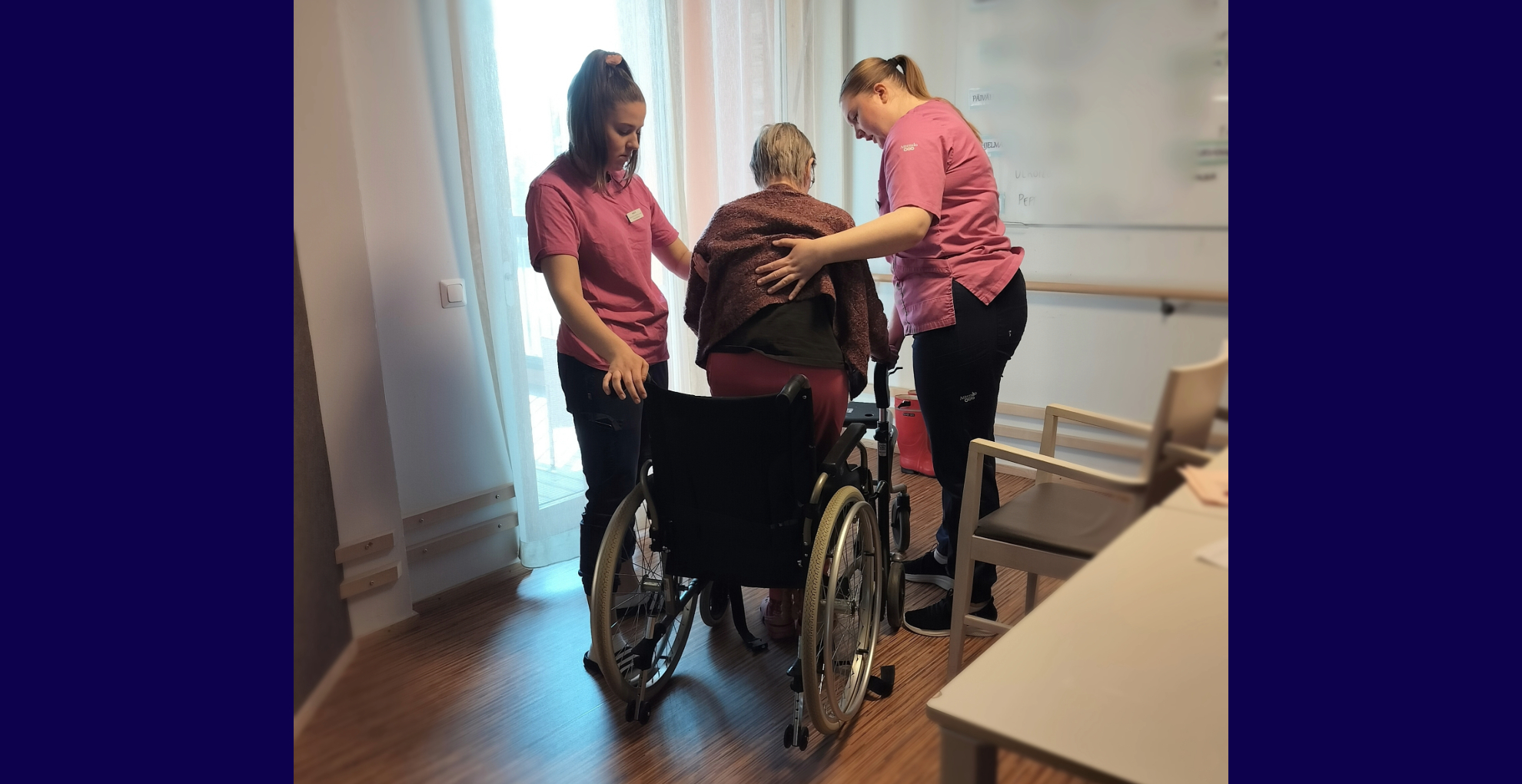 Kaksi hoitajaa avustaa pyörätuolissa olevaa vanhempaa henkilöä hoivakodissa.