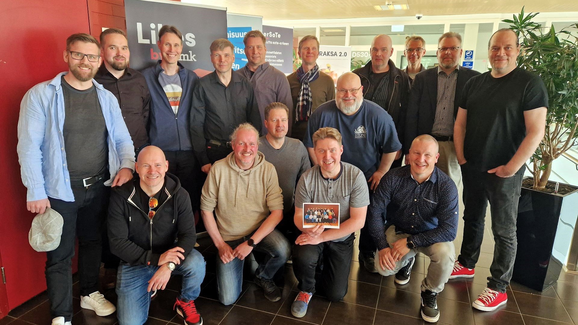 16 miestä hymyilee iloisesti kohti kameraa. Yhdellä on kädessä vanha luokkakuva.