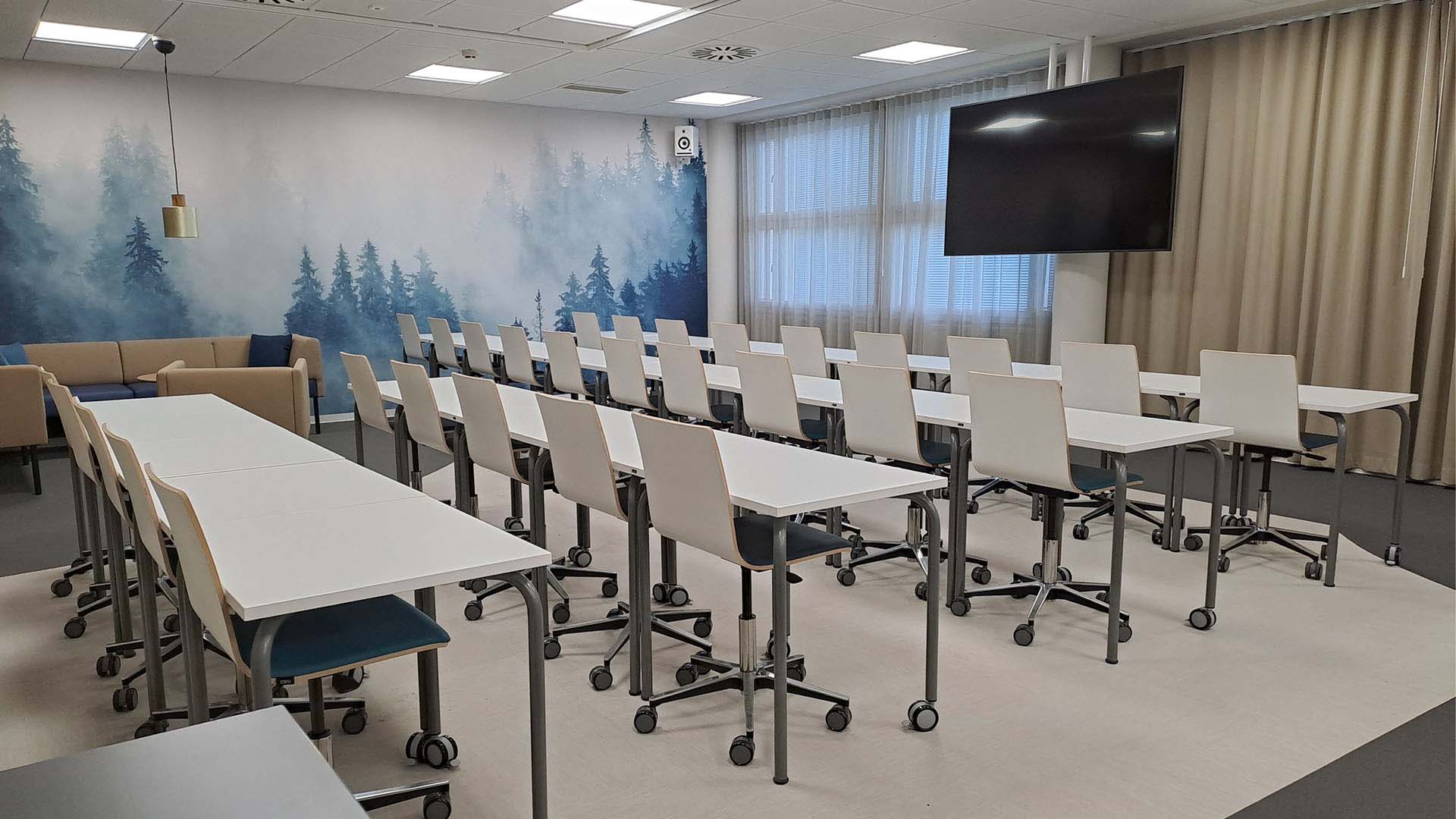 Takaseinällä sinisävyinen metsämaisematapetti ja sininen sohvaryhmä, keskellä valkoisia pulpetteja ja tuoleja riveissä, ikkunaseinustalla näyttö