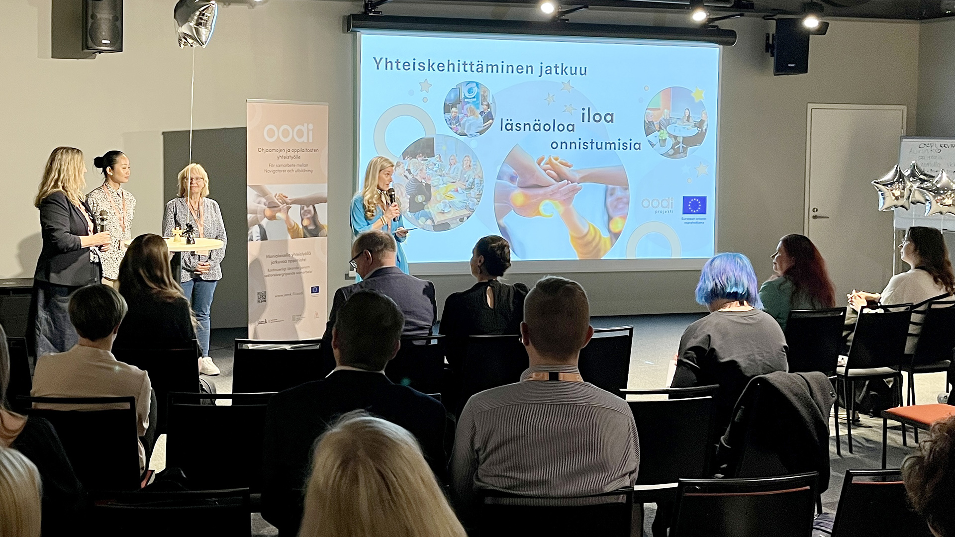 Tapahtuman yleisö istumassa tunnelmallisessa salissa ja salin edustalla neljä henkilöä esiintymässä.