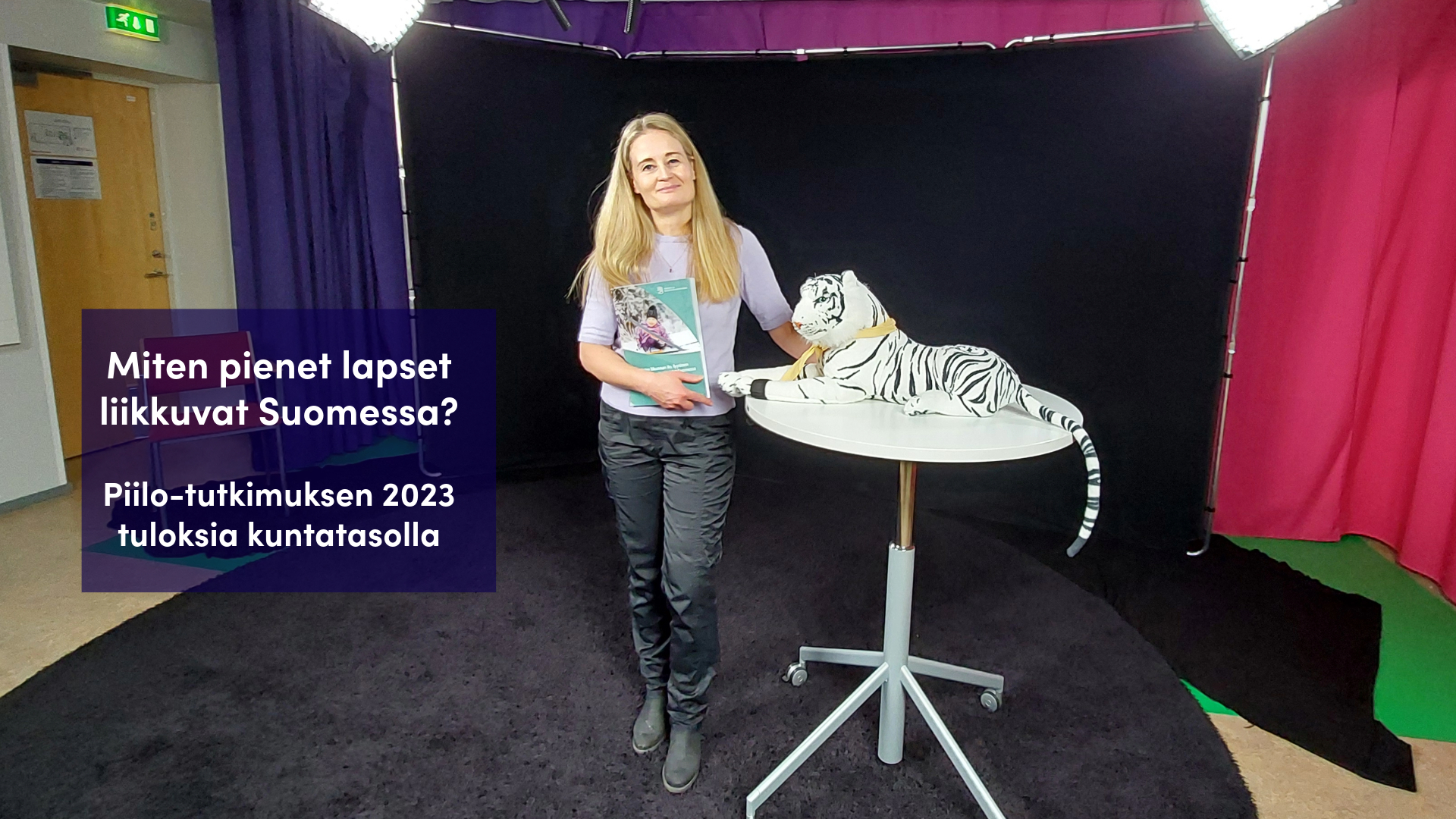 Anette Mehtälä studiossa Piilo-raportti kädessä ja pöydällä pehmolelutiikeri.