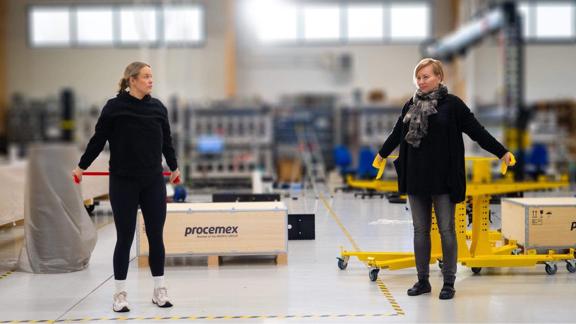 Kuvassa yrittäjä ja taukojumpan vetäjä Satu-Lotta Rytkönen sekä Procemex-yrityksen HR Specialist Riina Lestinen jumppaavat kuminauhan avulla Procemexin varastohallissa, tausta osittain sumennettu.