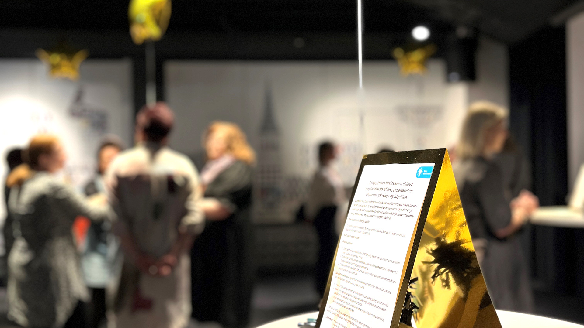 Epätarkka kuva tapahtuman työskentelyvaiheesta, jossa edustalla kultaisissa kehyksissä työskentelyn materiaalia.