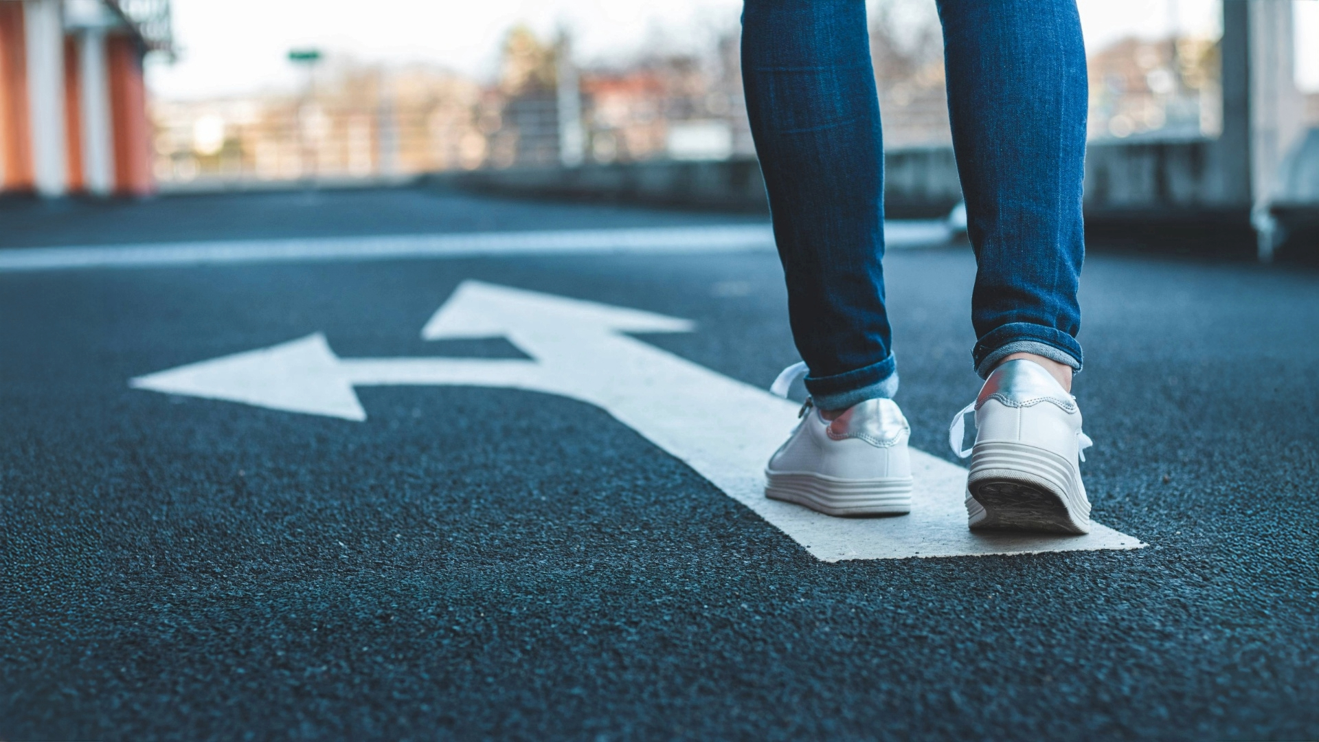 Farkkulahkeiset jalat seisovat asvalttiin maalatun nuolen päällä. Nuolen yksi kärki osoittaa suoraan, toinen kääntyy vasemmalle.