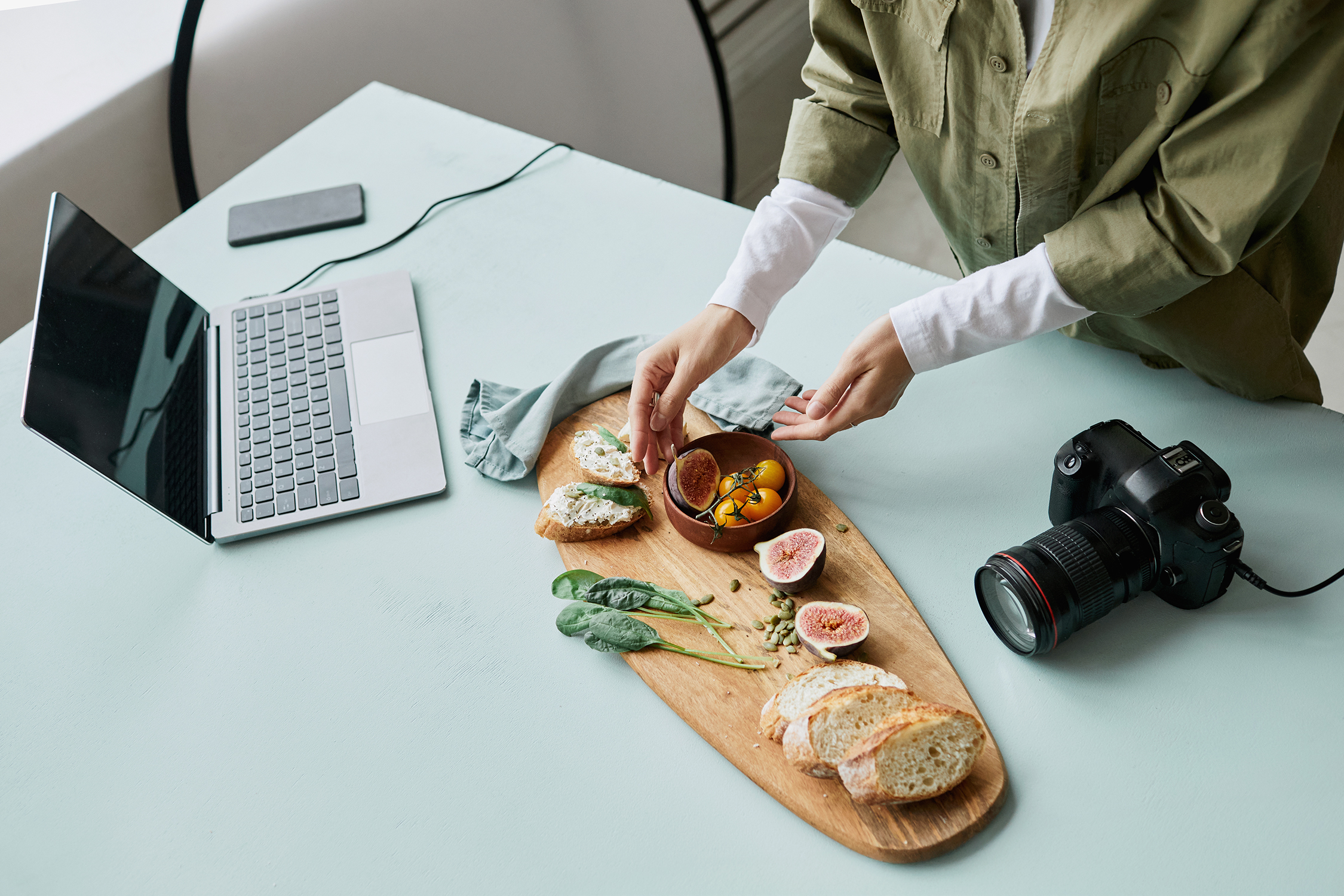 Henkilö asettelee ruokaa pöydän keskellä olevan leikkuulaudan päälle aikeissaan ottaa siitä kuvan vieressä olevalla kamerallaan