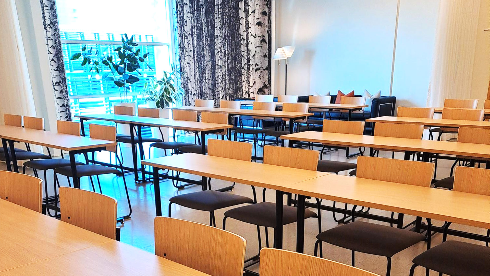 Takavasemmalla ikkunan edessä iso viherkasvi, koivutapettia ja iso valkea lamppu. Rivissä puisia pitkiä pöytiä ja pehmustettuja tuoleja