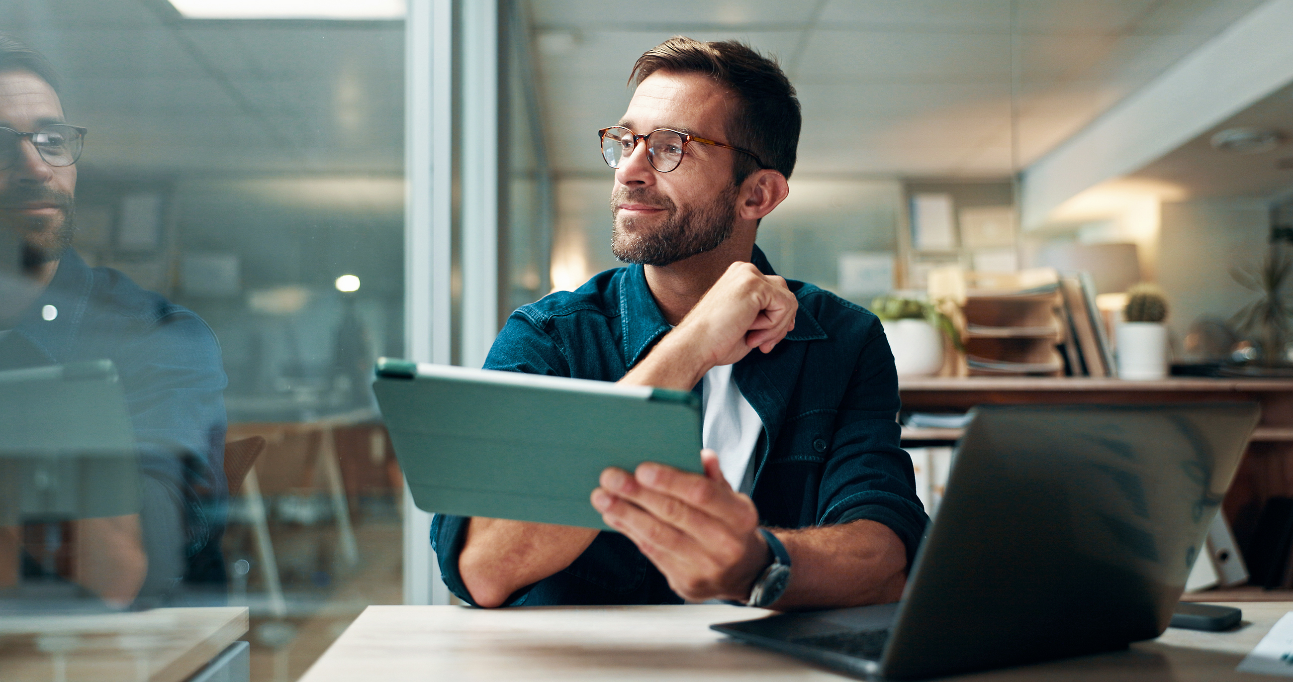 Man with eyeglasses sitting in an office holding a tablet and looking outside