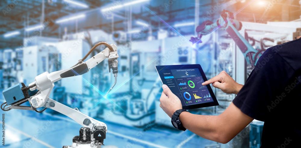 Henkilö tarkastelee tabletilla suorituskykydataa automatisoidussa teollisuusympäristössä, jossa näkyy robottikäsi ja muuta tuotantolaitteistoa.